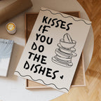Notebook with 'Kisses if you do the dishes' text and illustration on a wooden surface.