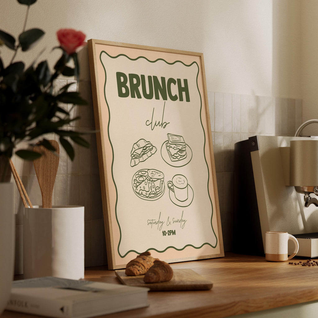 Framed poster with 'Brunch Club' text and food illustrations on a wooden surface with a plant and coffee maker in the background.
