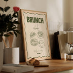 Framed poster with 'Brunch Club' text and food illustrations on a wooden surface with a plant and coffee maker in the background.