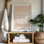 Framed artwork with 'So Fresh and So Clean' text on a wooden shelf with towels and a plant.