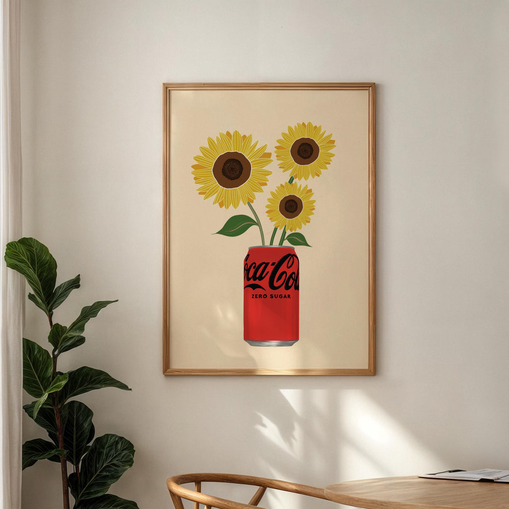 Framed artwork of sunflowers emerging from a Coca-Cola can on a wall with a plant and table in the foreground.