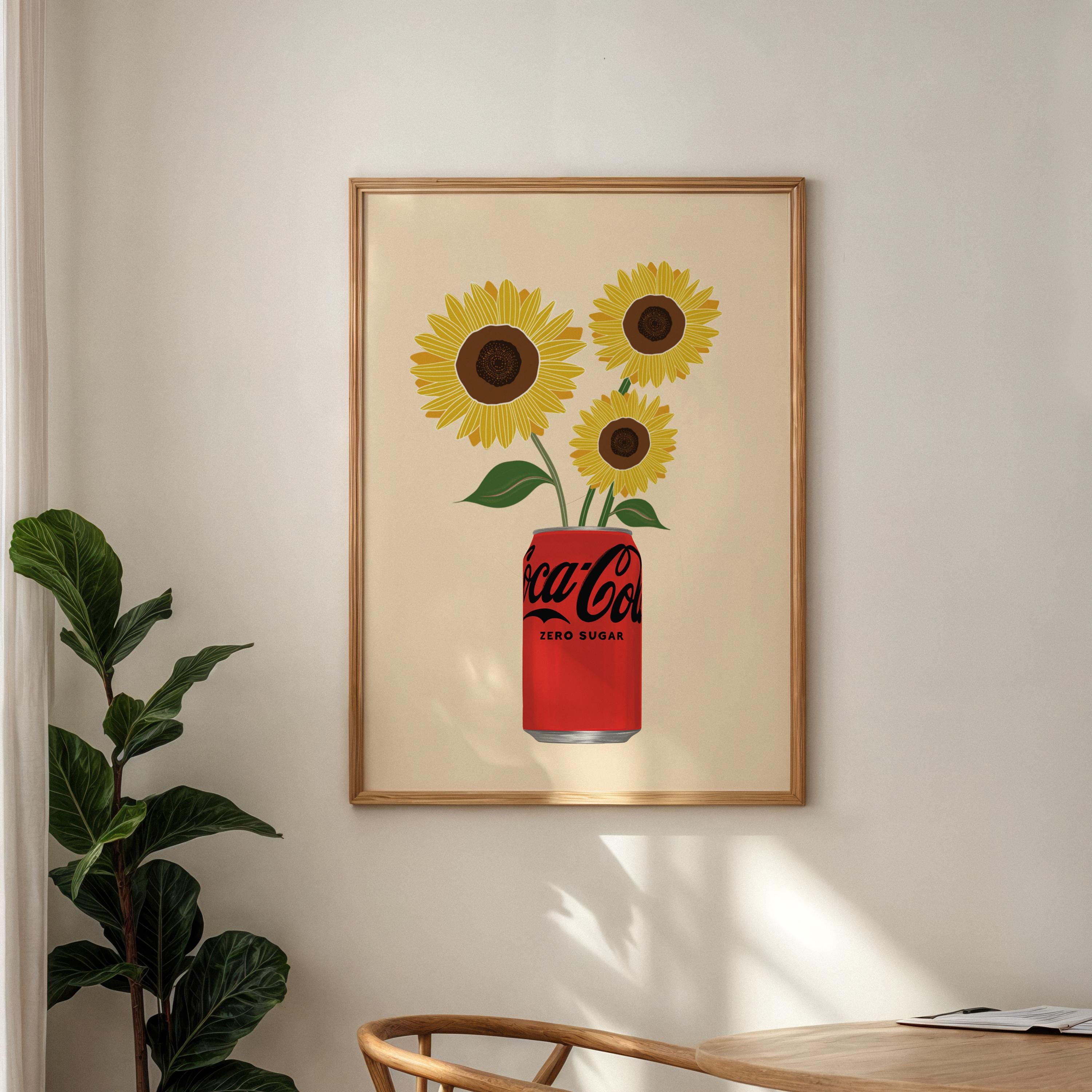 Framed artwork of sunflowers emerging from a Coca-Cola can on a wall with a plant and table in the foreground.