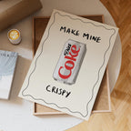 Notebook with 'Make Mine Diet Coke Crispy' text and illustration on a wooden surface.