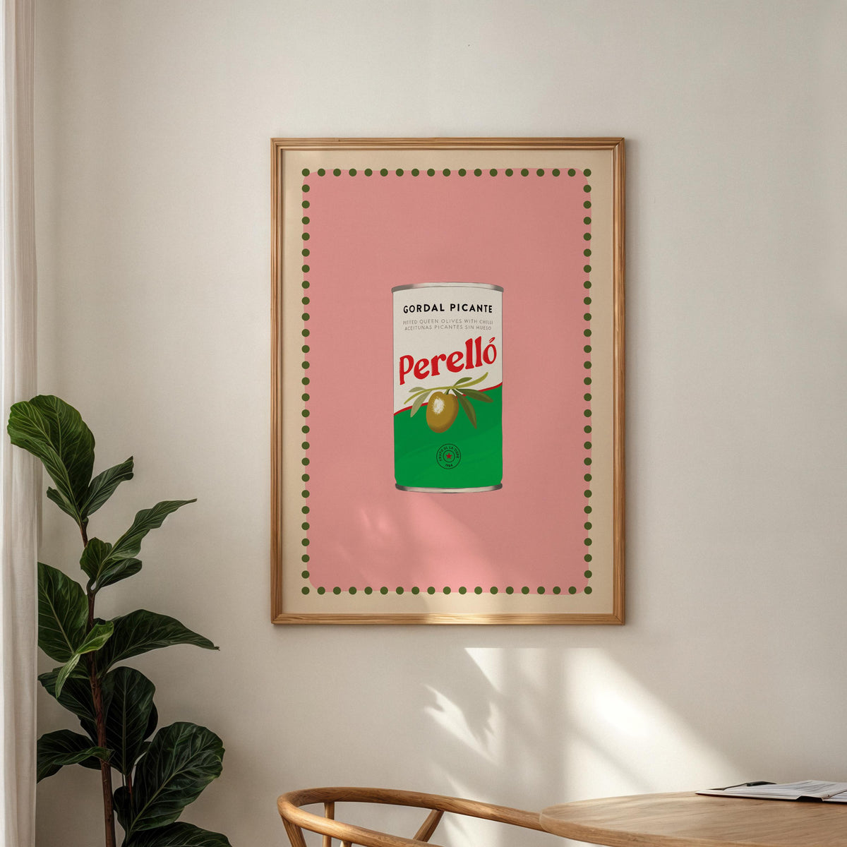 Framed artwork of a Perello olive can on a wall with a plant and table in the foreground.