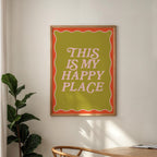 Framed poster with 'This is my happy place' text on a wall with a plant and table in the foreground.