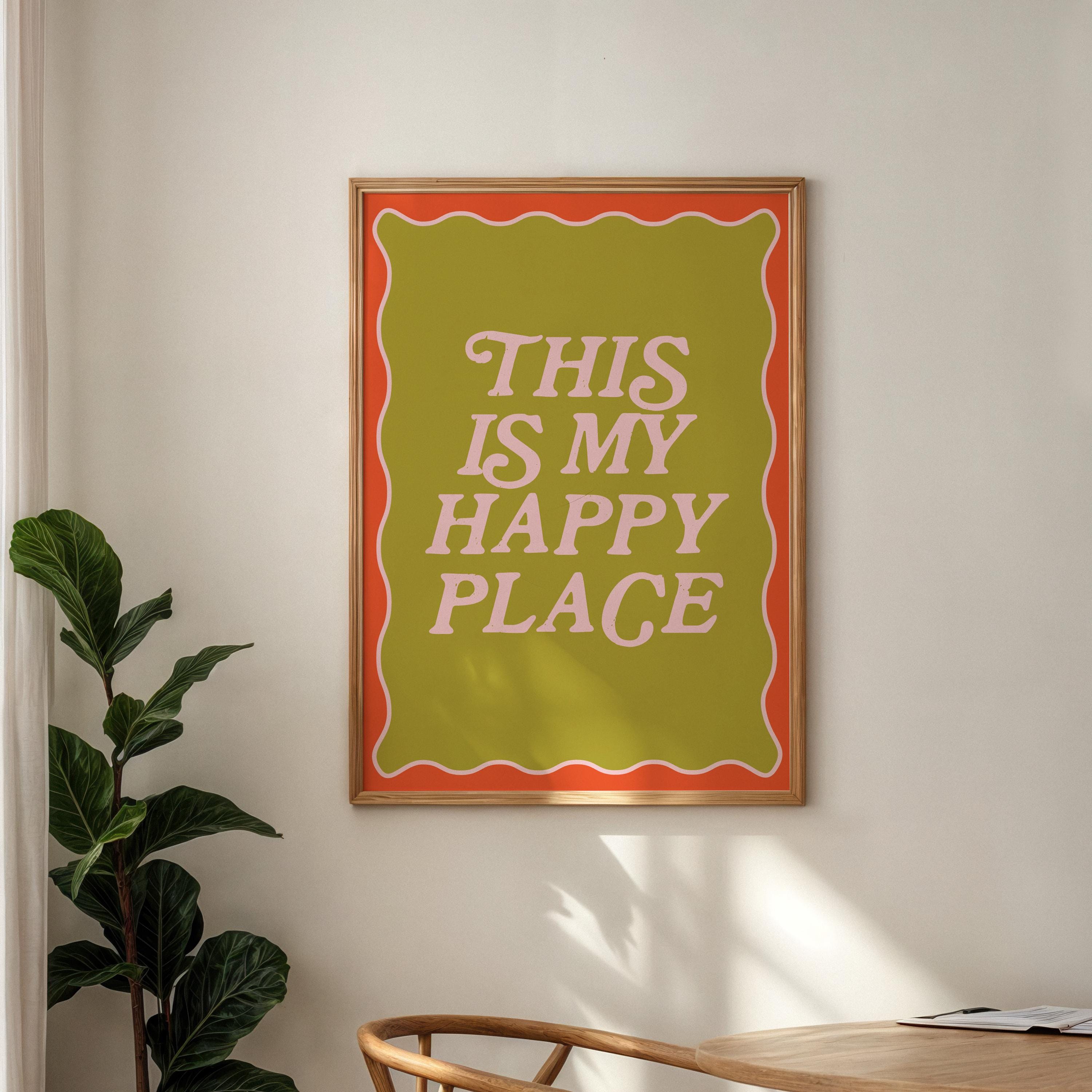 Framed poster with 'This is my happy place' text on a wall with a plant and table in the foreground.