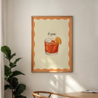 Framed artwork of a Negroni cocktail on a wall with a plant and table in the foreground.