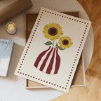 Print of sunflowers in a striped vase on a wooden surface with a cup of coffee and book.