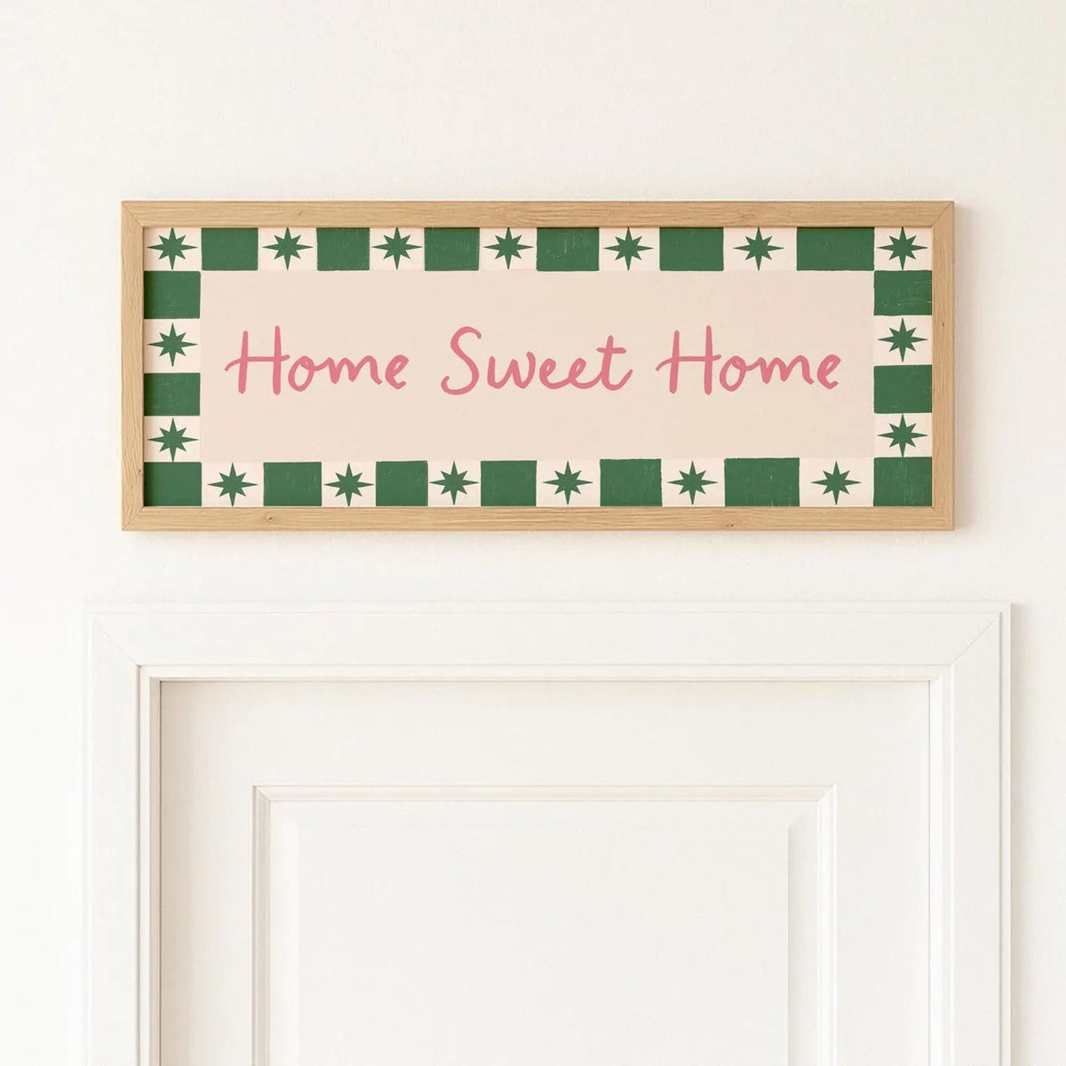 A rectangular framed wall sign with the phrase 'Home Sweet Home' in pink script, bordered by green and cream geometric patterns, hanging above a white door on a white wall.