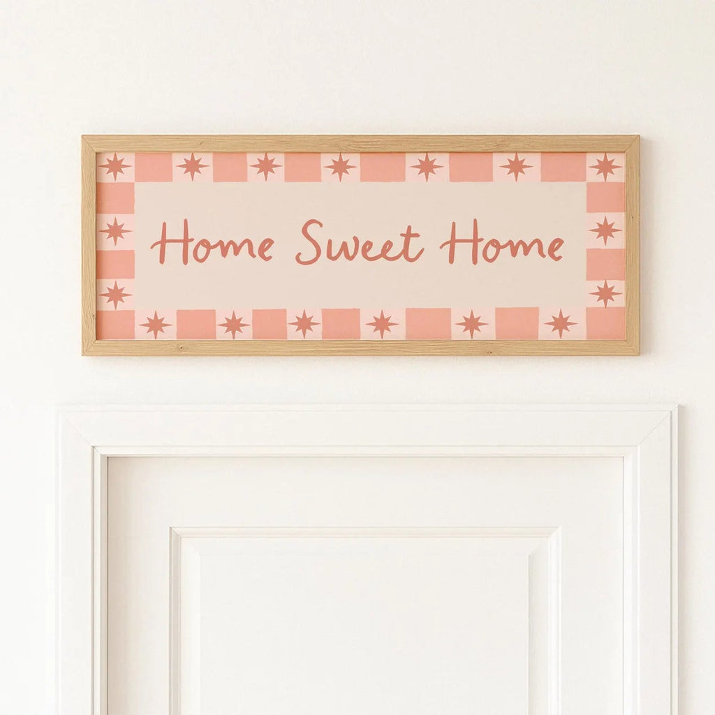 A rectangular wooden sign with the phrase 'Home Sweet Home' in cursive, decorated with a pink and peach gingham border and star motifs, mounted on a white wall above a white door frame.