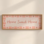 Framed decorative sign with 'Home Sweet Home' text on a checkered pattern against a beige wall.