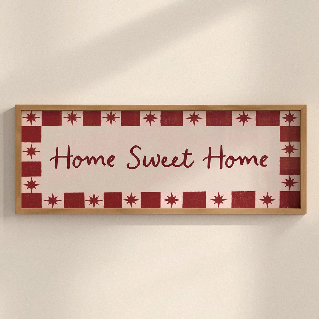A rectangular framed wall sign with the words “Home Sweet Home” in dark red script, surrounded by a decorative border of red stars and red-and-white squares, mounted on a light beige wall.