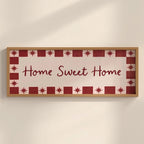 A rectangular framed wall sign with the words “Home Sweet Home” in dark red script, surrounded by a decorative border of red stars and red-and-white squares, mounted on a light beige wall.