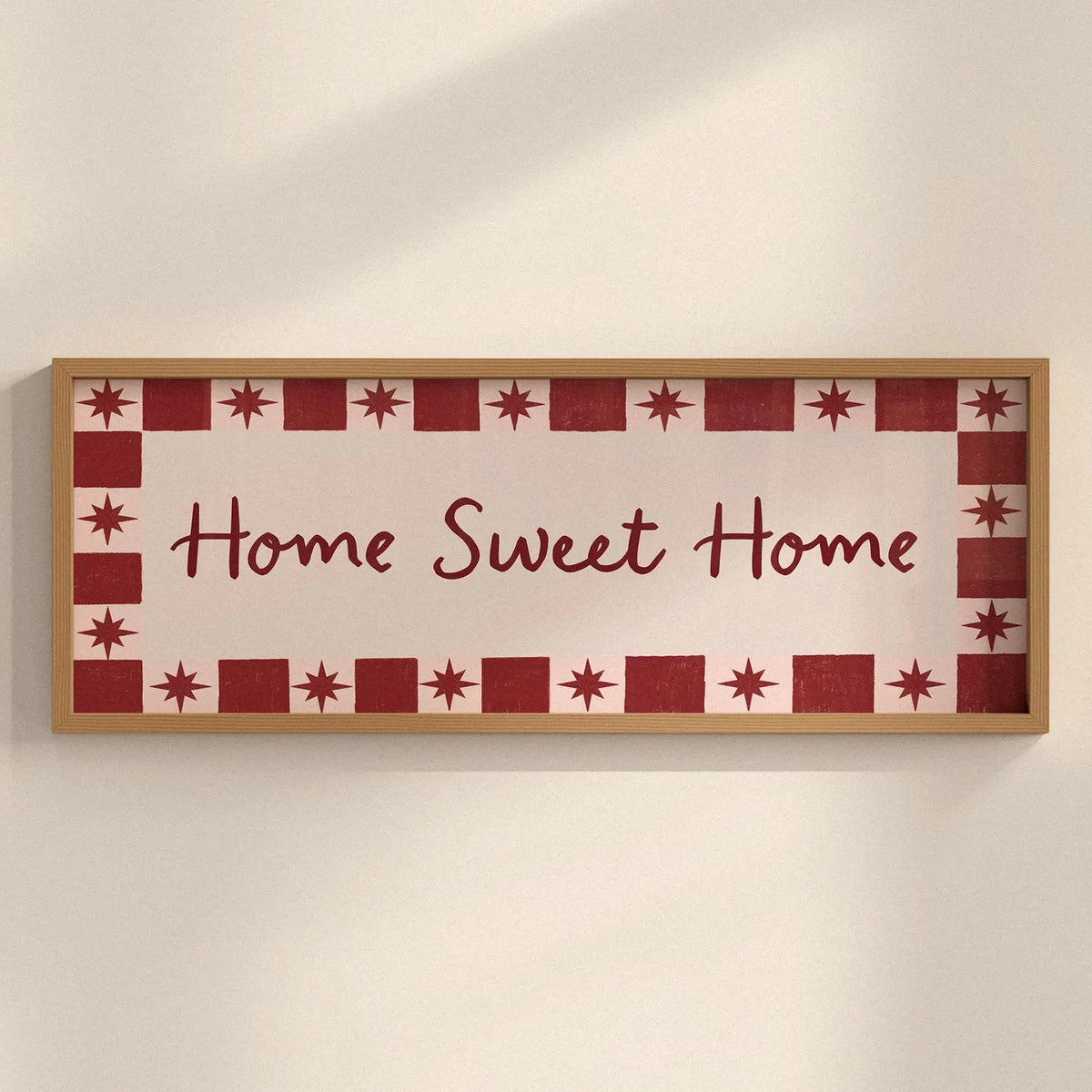 A rectangular framed wall sign with the words “Home Sweet Home” in dark red script, surrounded by a decorative border of red stars and red-and-white squares, mounted on a light beige wall.