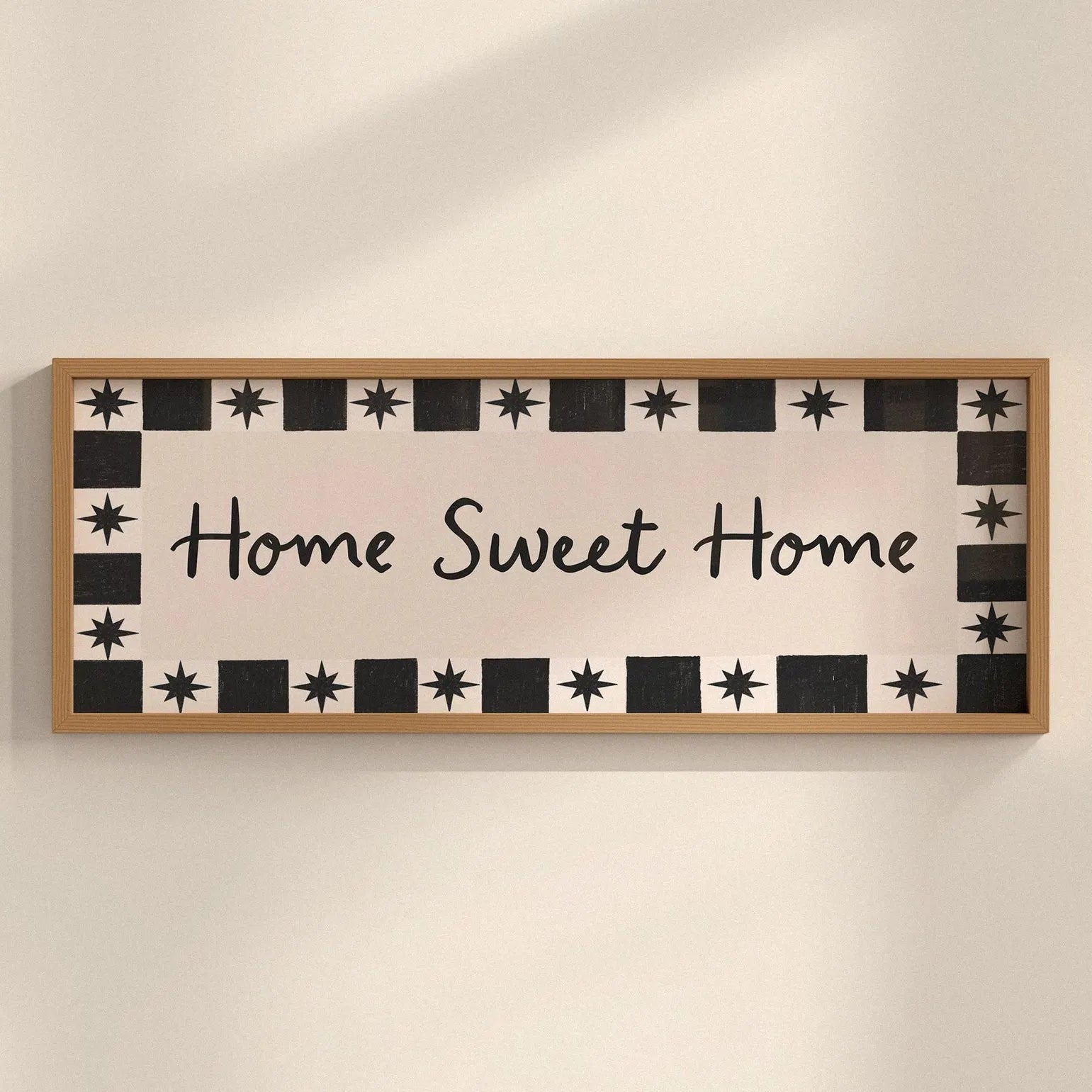 A rectangular wooden wall sign with the phrase 'Home Sweet Home' in black script, surrounded by a black-and-white geometric border of stars and squares, mounted on a warm beige wall.