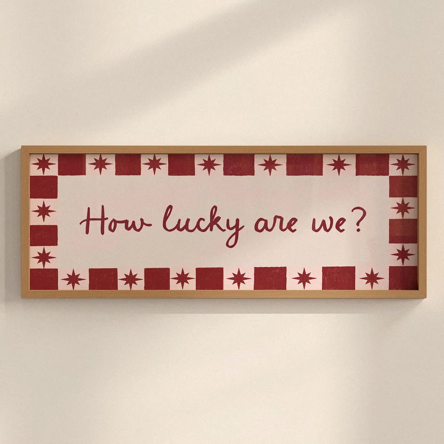A rectangular framed wall sign with the phrase “How lucky are we?” in red script, surrounded by a red and white star-and-square border. The sign is mounted on a plain off-white wall with soft shadows.