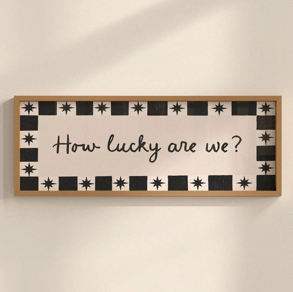 A rectangular wooden wall sign with a light natural wood frame, featuring a cream background with a black border made of alternating black stars and black squares, and the phrase 'How lucky are we?' written in black cursive script in the center.