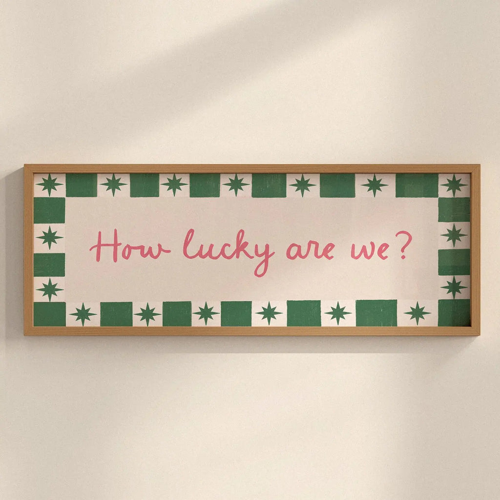 A rectangular framed wall sign with a light wood frame, featuring a cream background, a green and white patterned border, and the pink handwritten text 'How lucky are we?' mounted on a beige wall.