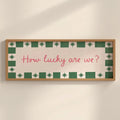 A rectangular framed wall sign with a light wood frame, featuring a cream background, a green and white patterned border, and the pink handwritten text 'How lucky are we?' mounted on a beige wall.