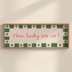 A rectangular framed wall sign with a light wood frame, featuring a cream background, a green and white patterned border, and the pink handwritten text 'How lucky are we?' mounted on a beige wall.