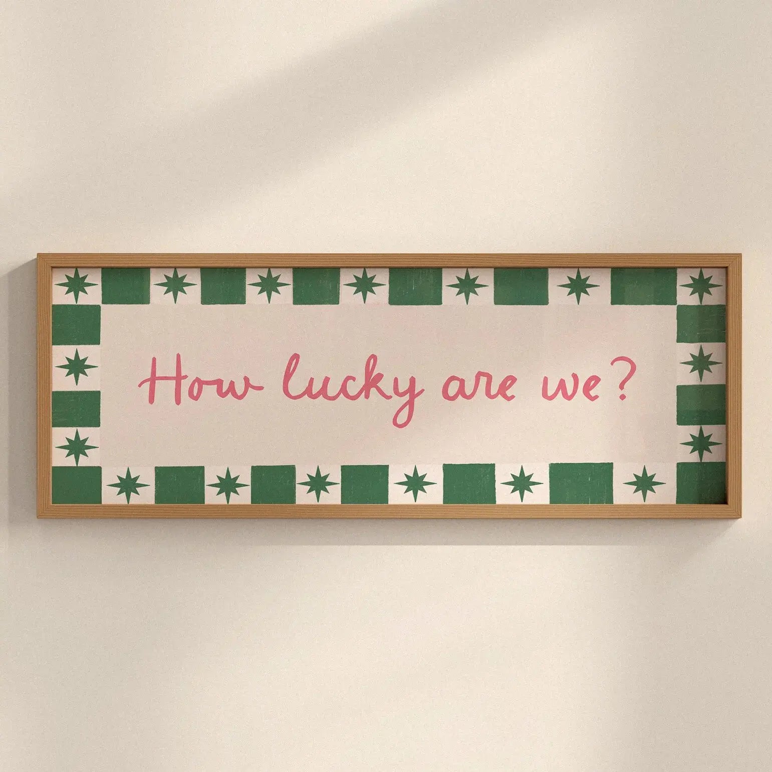 A rectangular framed wall sign with a light wood frame, featuring a cream background, a green and white patterned border, and the pink handwritten text 'How lucky are we?' mounted on a beige wall.