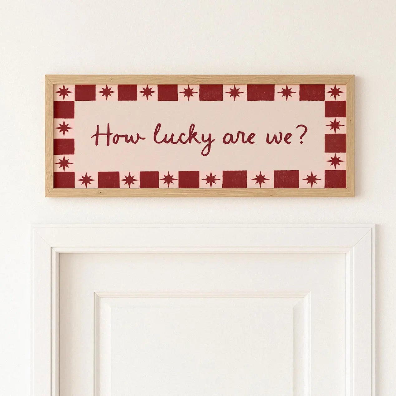 A rectangular framed wall sign with the phrase “How lucky are we?” in cursive, mounted above a white door on a white wall. The sign has a cream center, a red and cream checkered border with star motifs, and a light wood frame.