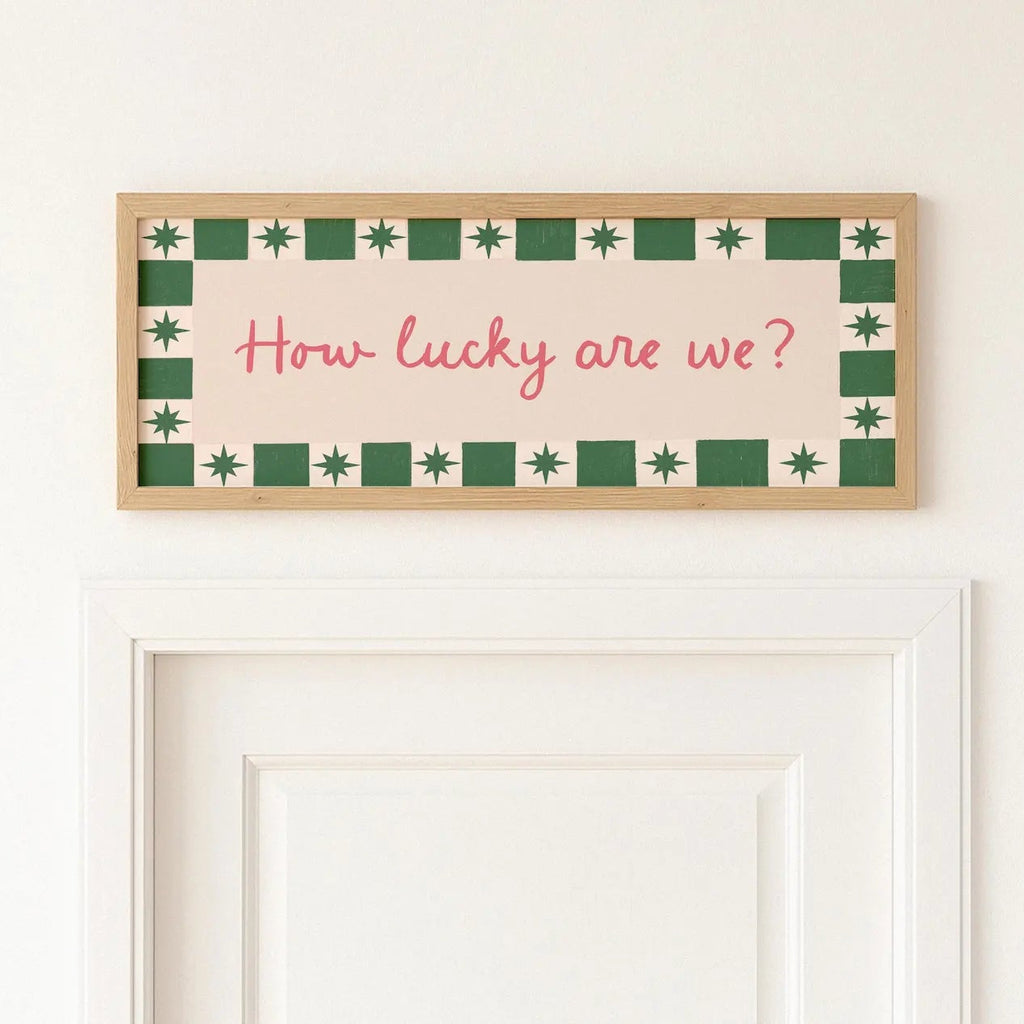 A rectangular framed wall sign with the phrase "How lucky are we?" in pink script, bordered by green and cream geometric patterns, hanging above a white door on a white wall