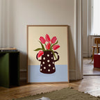 Framed artwork of a polka dot mug with red tulips on a beige and blue background, leaning against a wall.
