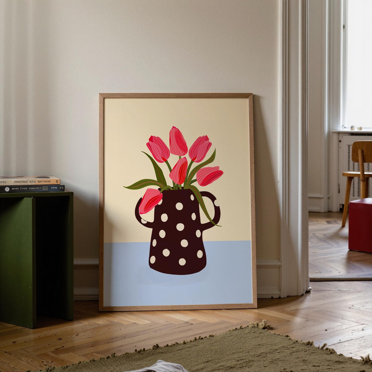 Framed artwork of a polka dot mug with red tulips on a beige and blue background, leaning against a wall.