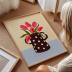 Framed artwork of a polka dot vase with pink flowers on a wooden floor.