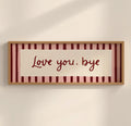 A framed horizontal wall art piece with the phrase 'Love you, bye' in dark red script over a cream center panel, set against pink and burgundy vertical stripes, in a light wood frame on a warm off-white wall.