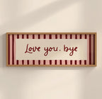 A framed horizontal wall art piece with the phrase 'Love you, bye' in dark red script over a cream center panel, set against pink and burgundy vertical stripes, in a light wood frame on a warm off-white wall.