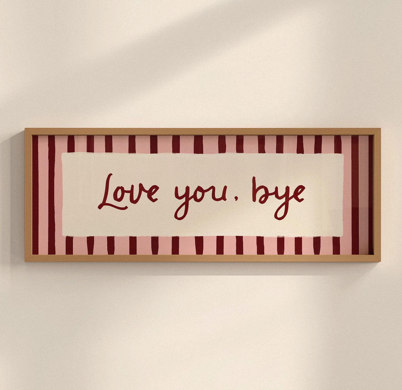A framed horizontal wall art piece with the phrase 'Love you, bye' in dark red script over a cream center panel, set against pink and burgundy vertical stripes, in a light wood frame on a warm off-white wall.