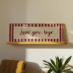A rectangular framed wall sign with the phrase 'Love you, bye' in dark red script, set against a striped background of dark red and pale pink with a light wood frame. The sign is displayed on a light wood floating shelf against a warm beige wall, with a mustard yellow chair and a green potted plant visible in the foreground.