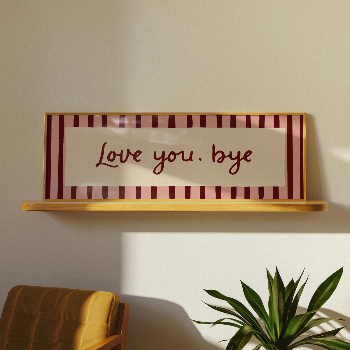 A rectangular framed wall sign with the phrase 'Love you, bye' in dark red script, set against a striped background of dark red and pale pink with a light wood frame. The sign is displayed on a light wood floating shelf against a warm beige wall, with a mustard yellow chair and a green potted plant visible in the foreground.