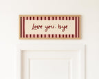 A rectangular framed wall sign with the words "Love you, bye" in dark red script, set against a cream center panel and a border of alternating cream and dark red stripes. The sign is mounted on a white wall above a white door.