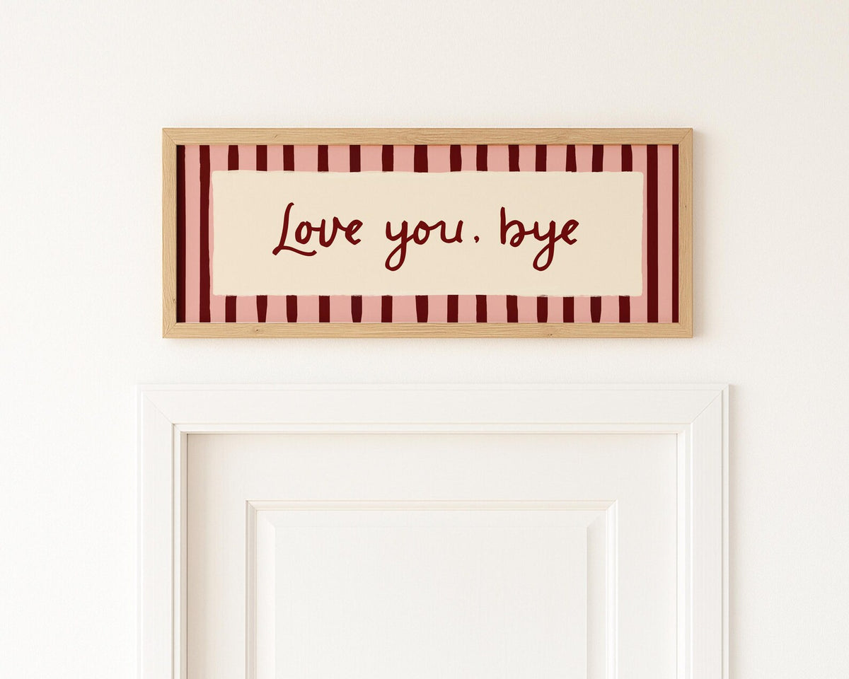 A rectangular framed wall sign with the words "Love you, bye" in dark red script, set against a cream center panel and a border of alternating cream and dark red stripes. The sign is mounted on a white wall above a white door.