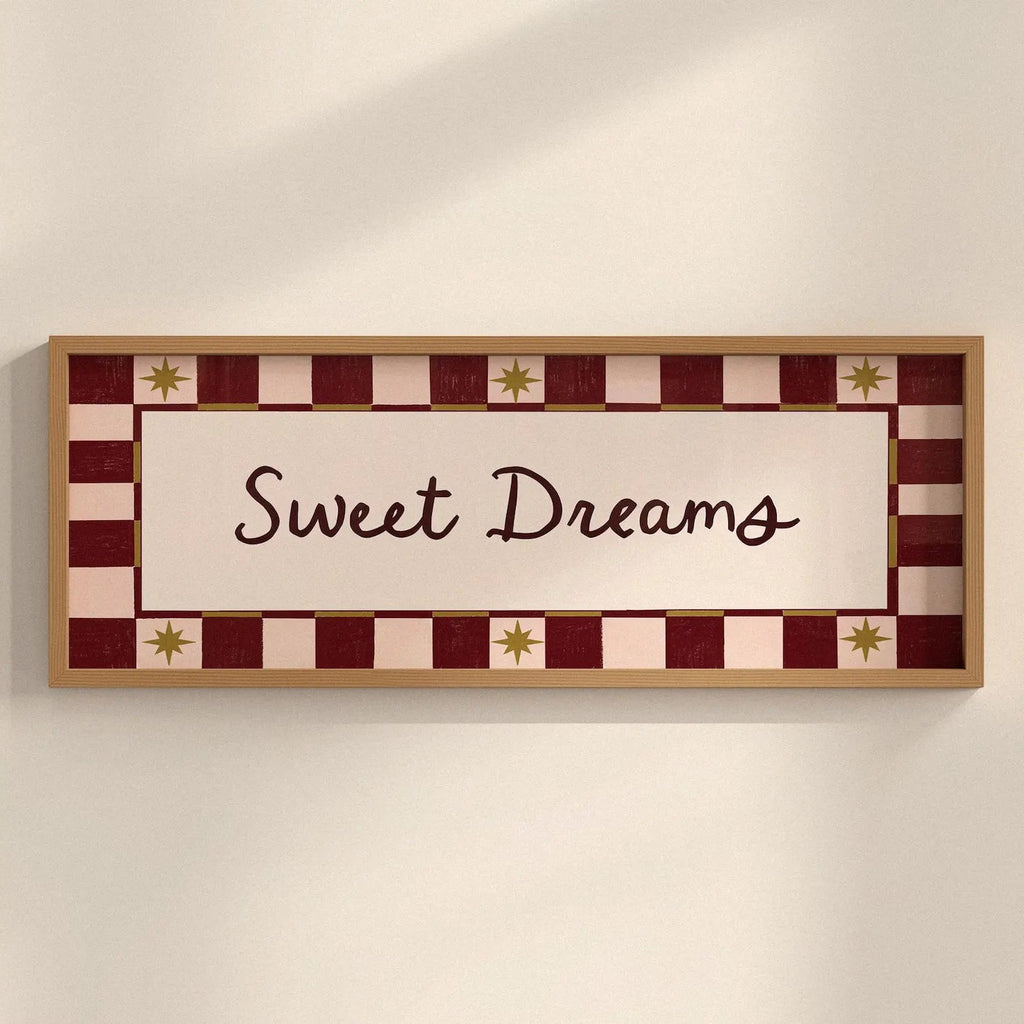 A rectangular framed wall sign with the words 'Sweet Dreams' in dark script on a cream center panel, surrounded by a red and cream checkered border with small gold star motifs, shown hanging on a light beige wall.
