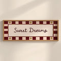 A rectangular framed wall sign with the words 'Sweet Dreams' in dark script on a cream center panel, surrounded by a red and cream checkered border with small gold star motifs, shown hanging on a light beige wall.
