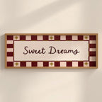 A rectangular framed wall sign with the words 'Sweet Dreams' in dark script on a cream center panel, surrounded by a red and cream checkered border with small gold star motifs, shown hanging on a light beige wall.