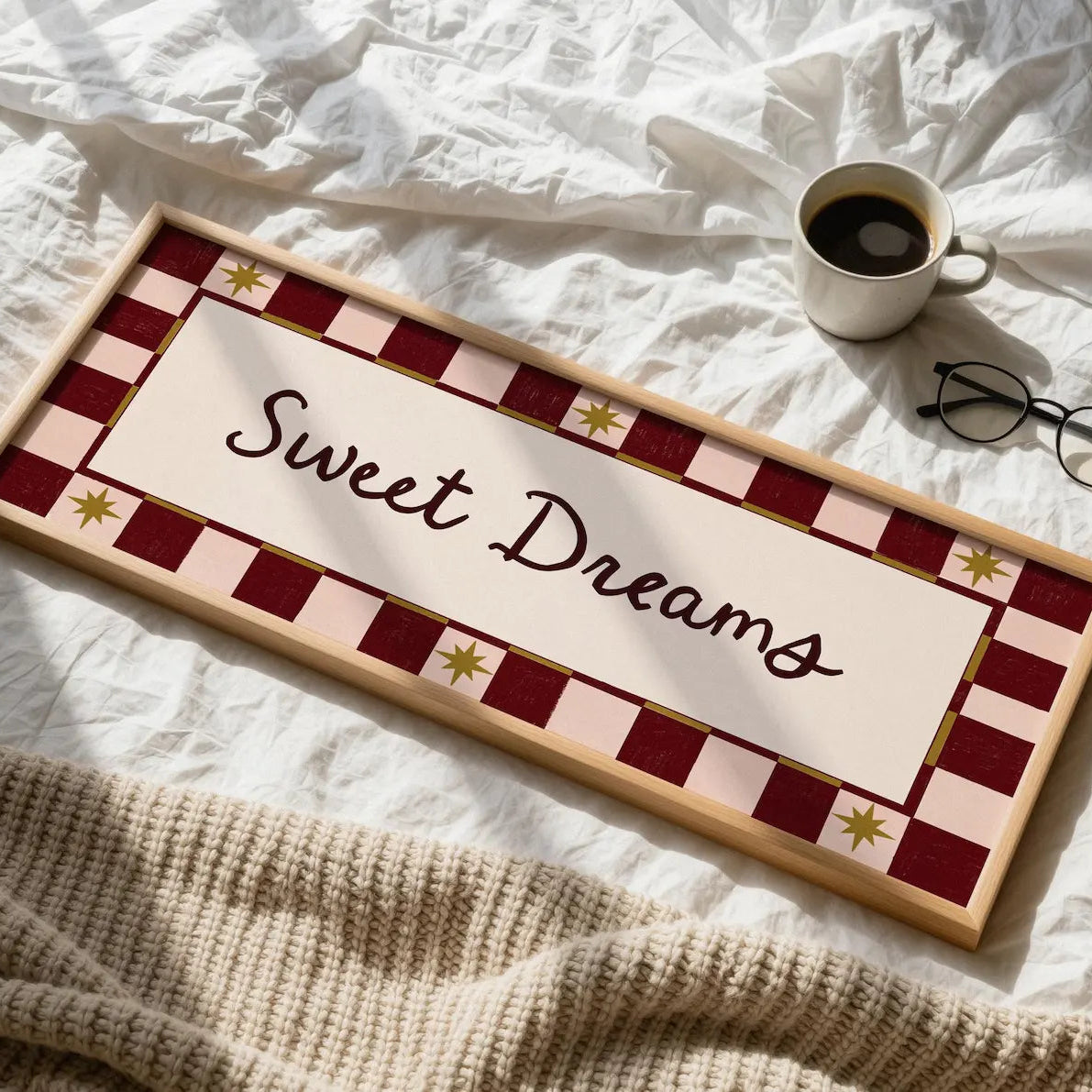 A rectangular decorative sign reading “Sweet Dreams” in black script, placed on a white bed with a cup of coffee, eyeglasses, and a beige knit blanket nearby. The sign has a cream center panel with a red-and-white check border and small gold star accents.