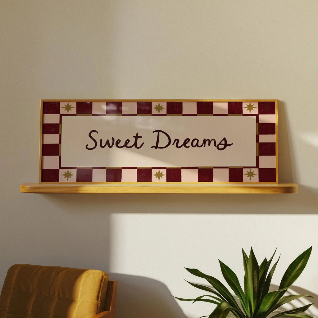 A decorative wall sign reading 'Sweet Dreams' in dark script, mounted on a light wood shelf against a beige wall. The sign has a cream center panel surrounded by a burgundy and cream checkered border with small gold star motifs.