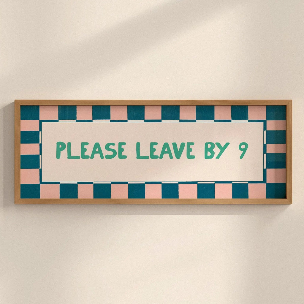 A rectangular framed sign with a light wood frame, featuring a pink and teal checkered border and the text 'PLEASE LEAVE BY 9' in teal on a cream background, mounted on a light beige wall.