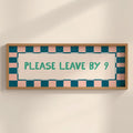 A rectangular framed sign with a light wood frame, featuring a pink and teal checkered border and the text 'PLEASE LEAVE BY 9' in teal on a cream background, mounted on a light beige wall.