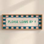 A rectangular framed sign with a light wood frame, featuring a pink and teal checkered border and the text 'PLEASE LEAVE BY 9' in teal on a cream background, mounted on a light beige wall.