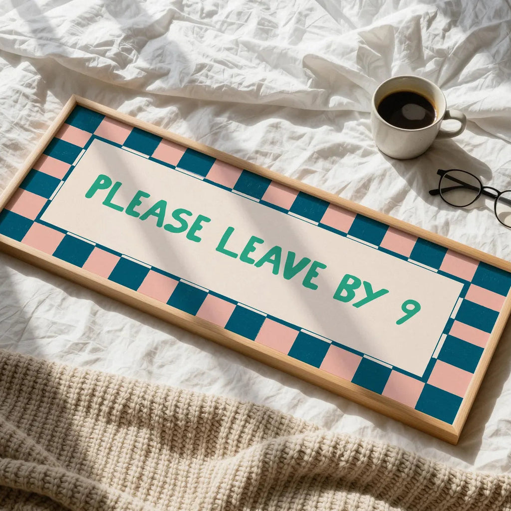 A rectangular wooden sign with a pastel pink and teal checkered border and the text 'PLEASE LEAVE BY 9' in teal, placed on white bedding with a mug, glasses, and a knitted blanket nearby.