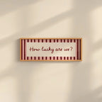 A rectangular framed wall sign with the phrase “How lucky are we?” in dark red script, set against a cream background with vertical dark red stripes and a light wood frame, mounted on a beige wall with soft diagonal shadows.