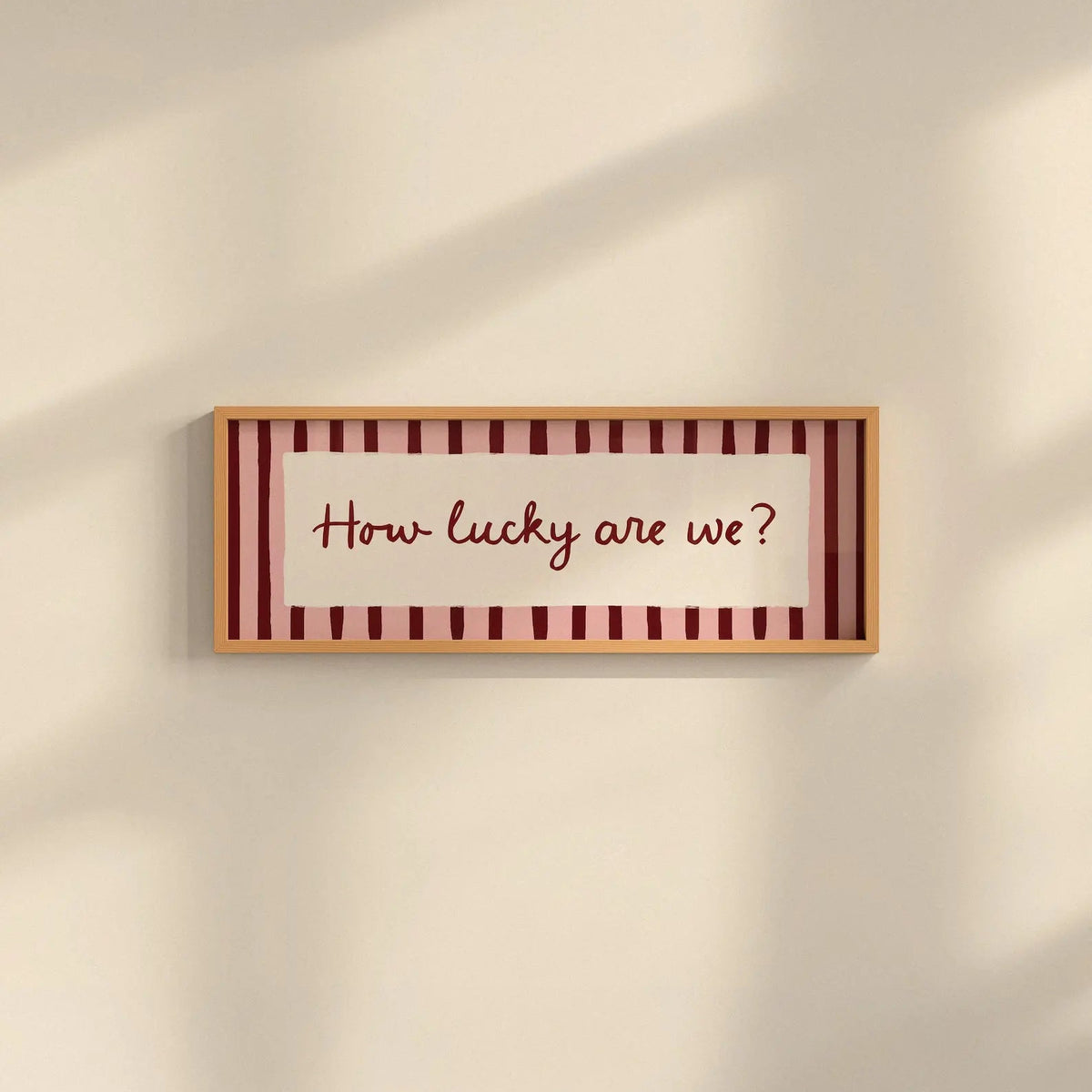 A rectangular framed wall sign with the phrase “How lucky are we?” in dark red script, set against a cream background with vertical dark red stripes and a light wood frame, mounted on a beige wall with soft diagonal shadows.