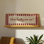 A framed wall sign with the phrase 'How lucky are we?' in dark red script, set against a cream center panel and burgundy-and-cream striped borders, mounted on a light wood shelf. The scene is styled in a room with a mustard chair and a green plant visible below.
