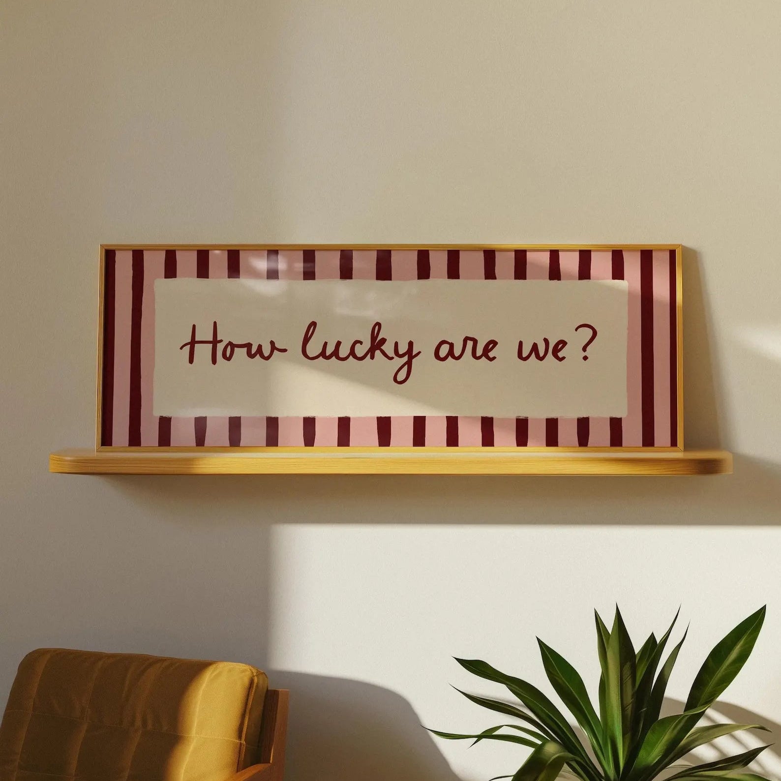 A framed wall sign with the phrase 'How lucky are we?' in dark red script, set against a cream center panel and burgundy-and-cream striped borders, mounted on a light wood shelf. The scene is styled in a room with a mustard chair and a green plant visible below.